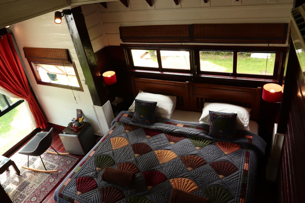 A bedroom with a large bed, wooden headboard, two bedside lamps, a window with a view of greenery, and a small table with a chair.