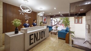 A hotel reception desk with a 'ENJOY' sign, two receptionists, a modern chandelier, and a seating area with a blue sofa. - Pergola Hotel Design (Da Nang)