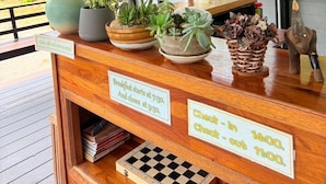 Een houten receptiebalie met bloempotten en een schaakbord. Borden geven de ontbijttijden en in- en uitchecktijden aan.