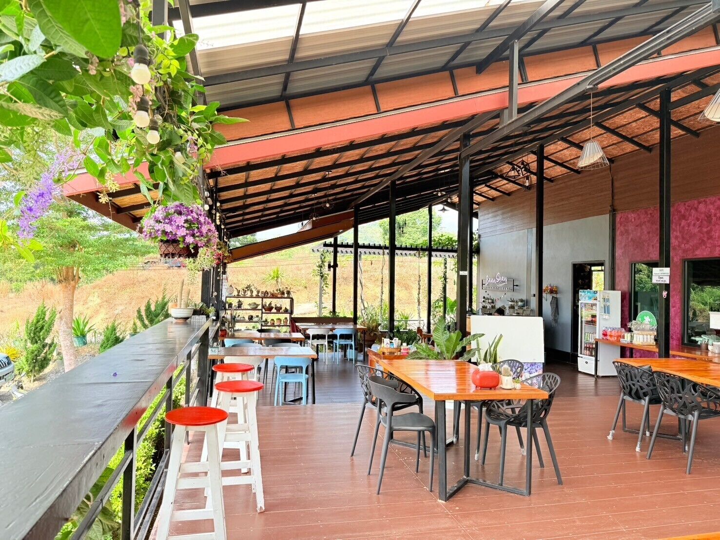 Un café en plein air avec des tables et des chaises en bois, entouré de plantes en pot et de fleurs suspendues.