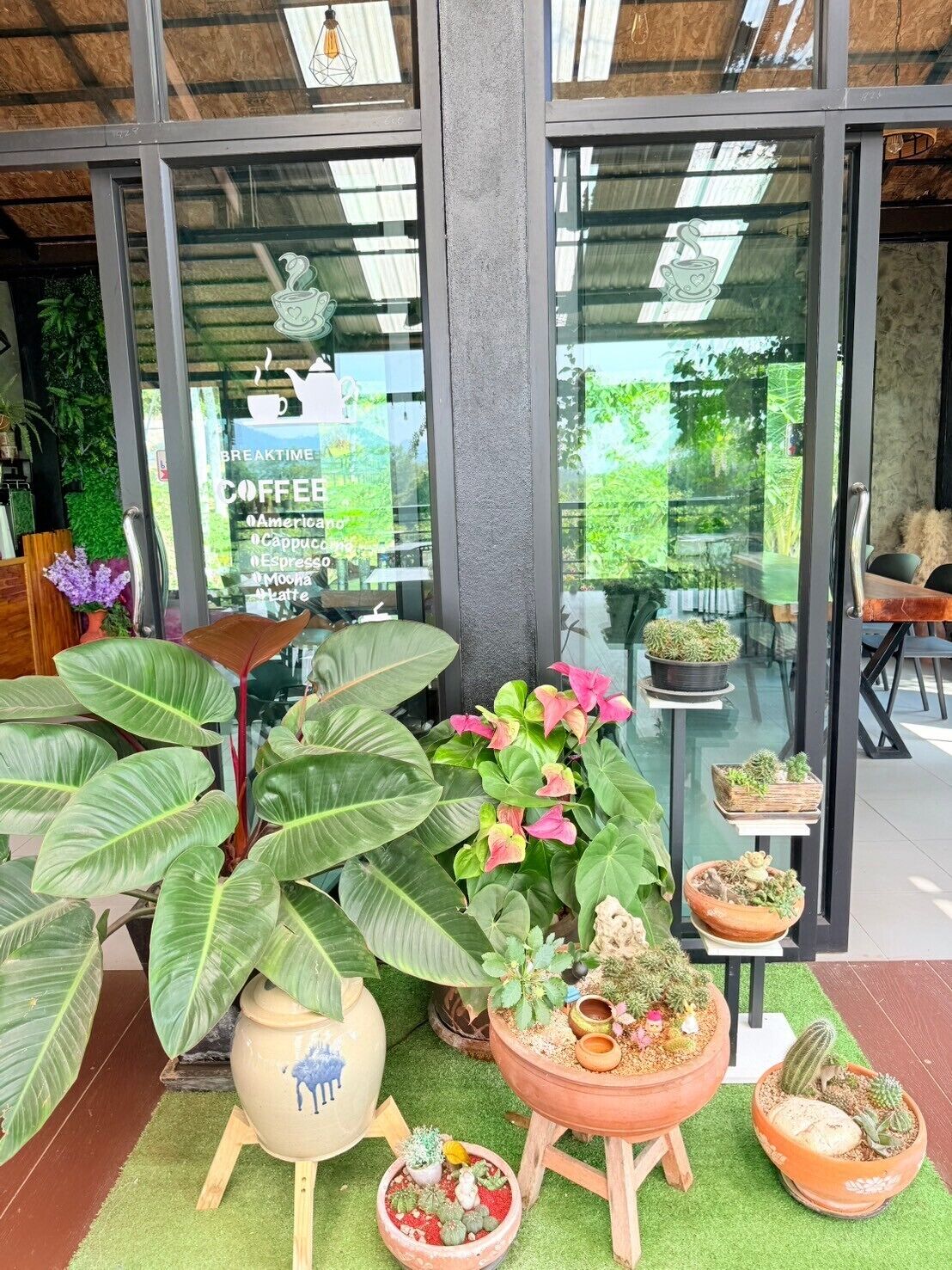 A coffee shop with large windows, various potted plants, and a sign listing coffee types.