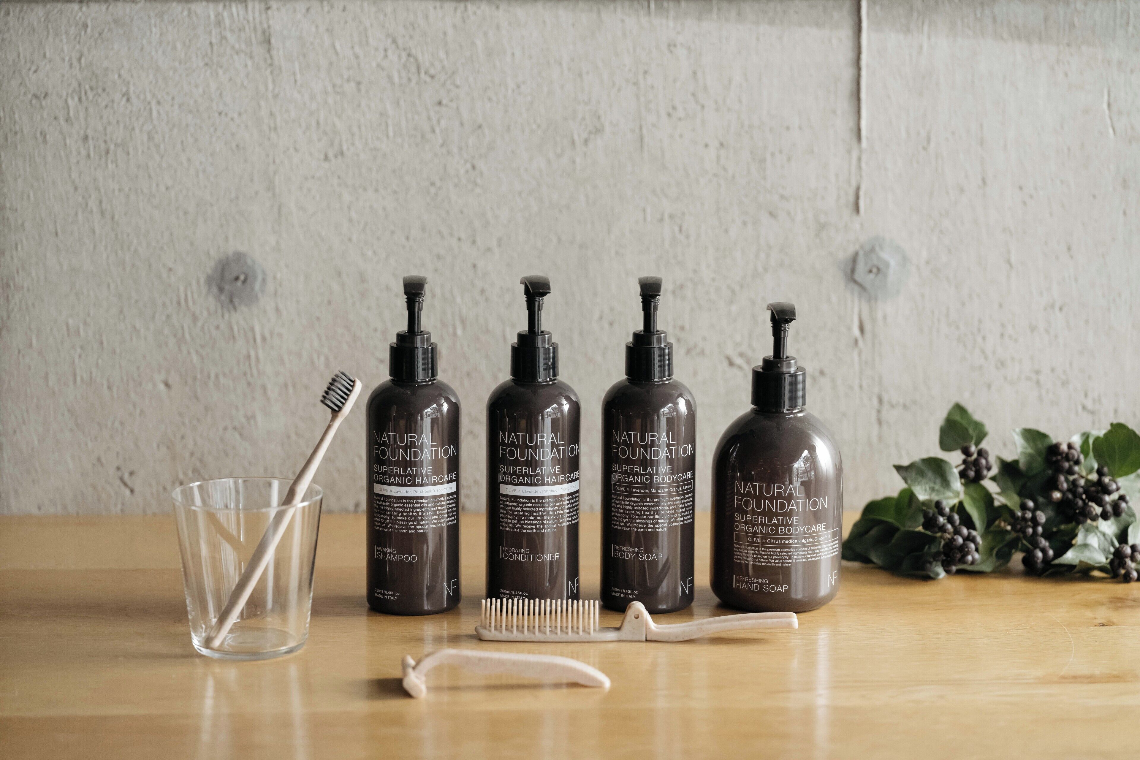 Four bottles of natural foundation superlative organic bodycare, a clear glass, and a white brush on a wooden surface.