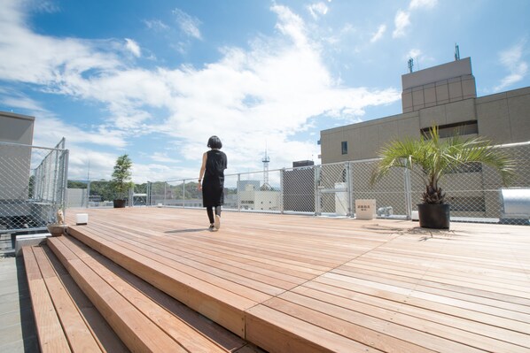 Una terraza de madera con escalones que conducen a un área en la azotea, rodeada de cercas de metal y edificios.