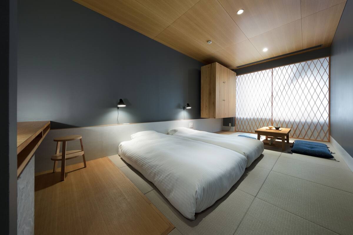 A modern bedroom with two beds, a wooden desk, a small stool, and a table with a bowl.