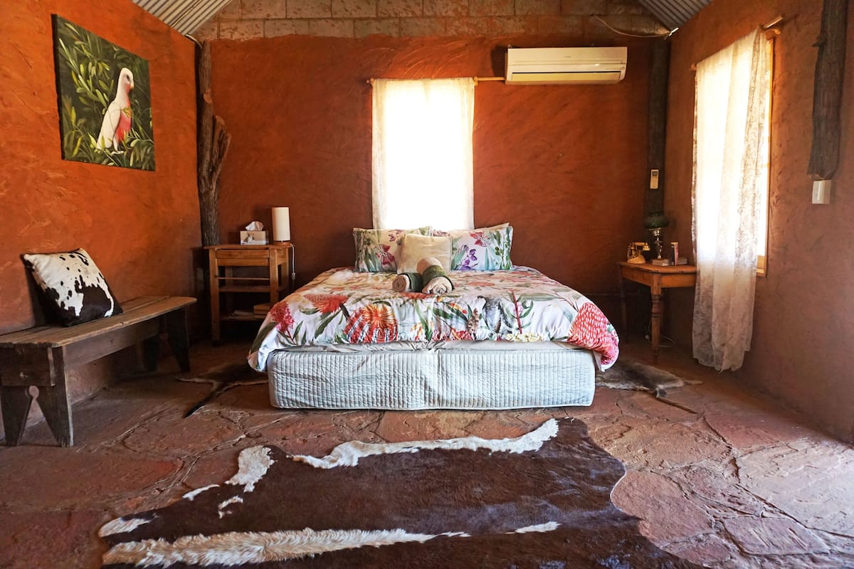 Ein rustikales Schlafzimmer mit Steinboden, einem Bett mit floralem Bettzeug, einer Holzbank und einer wandmontierten Klimaanlage.