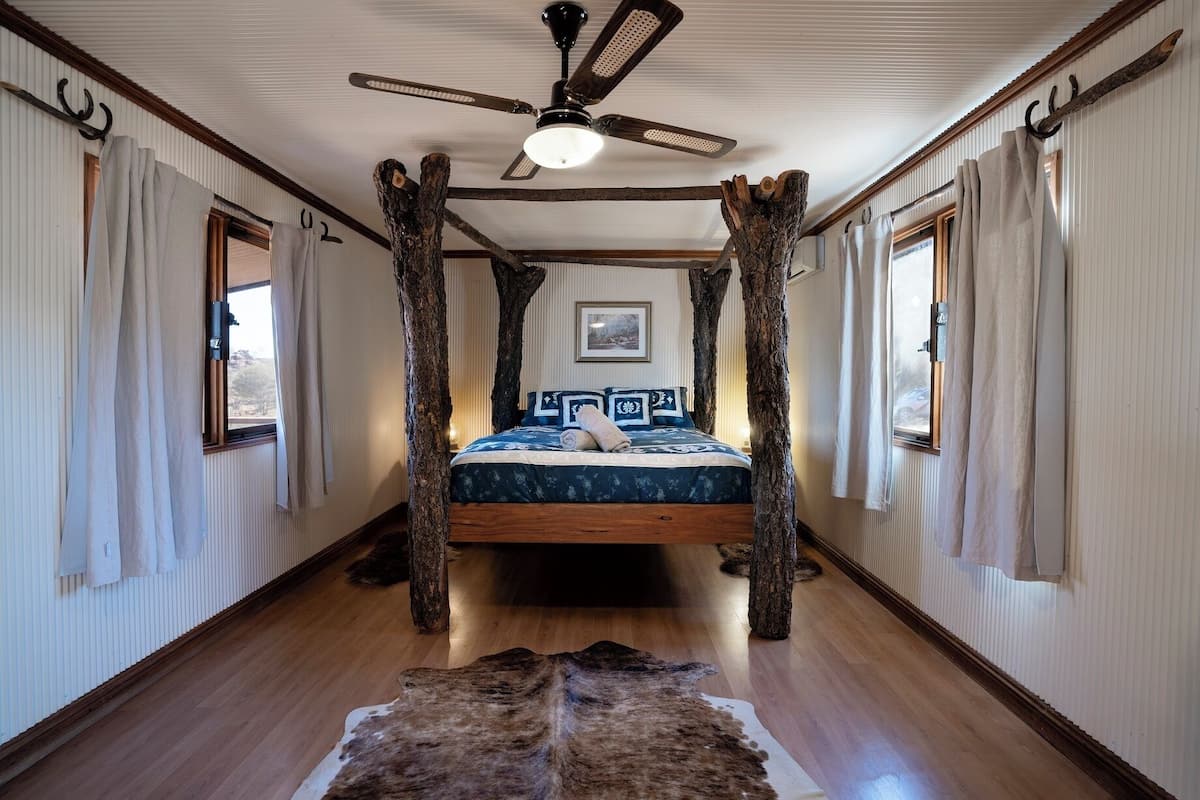 Ein Zimmer mit einem zentral platzierten Bett, Holzbalken, einem Deckenventilator und einem Fellteppich.