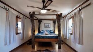Une chambre avec un lit au centre, des poutres en bois, un ventilateur de plafond et un tapis en fourrure.