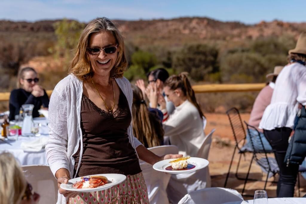 A person holding a plate with food at an outdoor dining area with tables and chairs, and a natural landscape in the background.