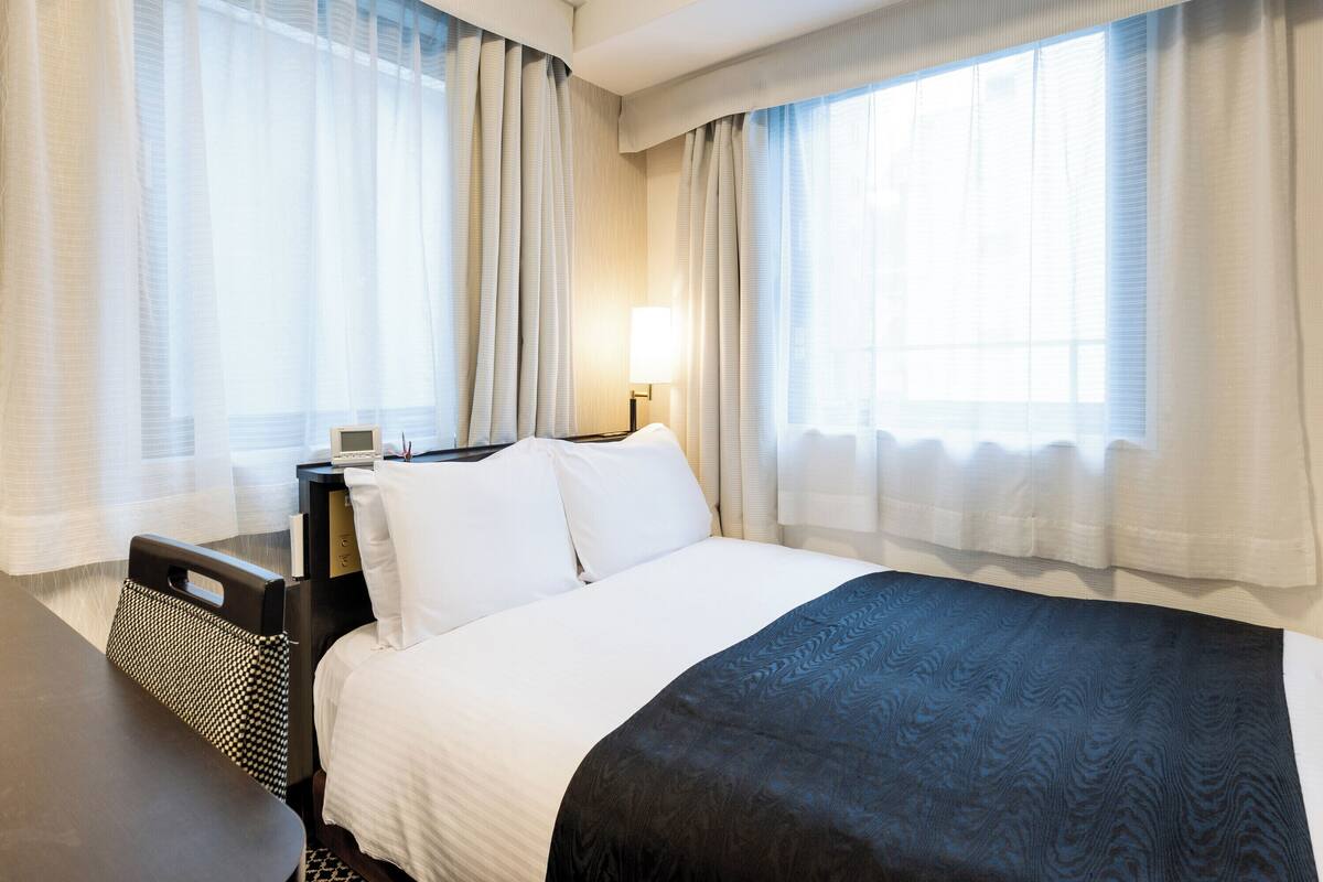 Een hotelkamer met een groot bed, een nachtkastje met een lamp en dunne gordijnen voor de ramen.