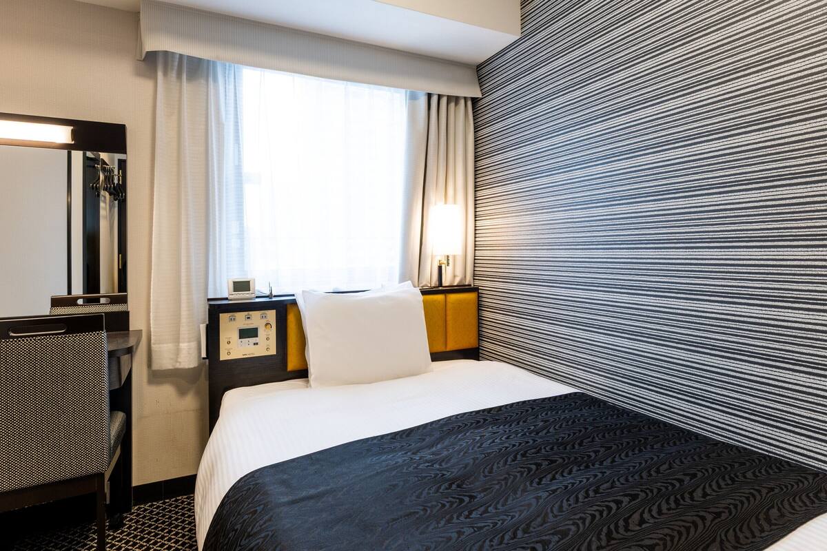 Een hotelkamer met een bed, een bureau, een stoel, een nachtkastje met lamp en een raam met gordijnen.