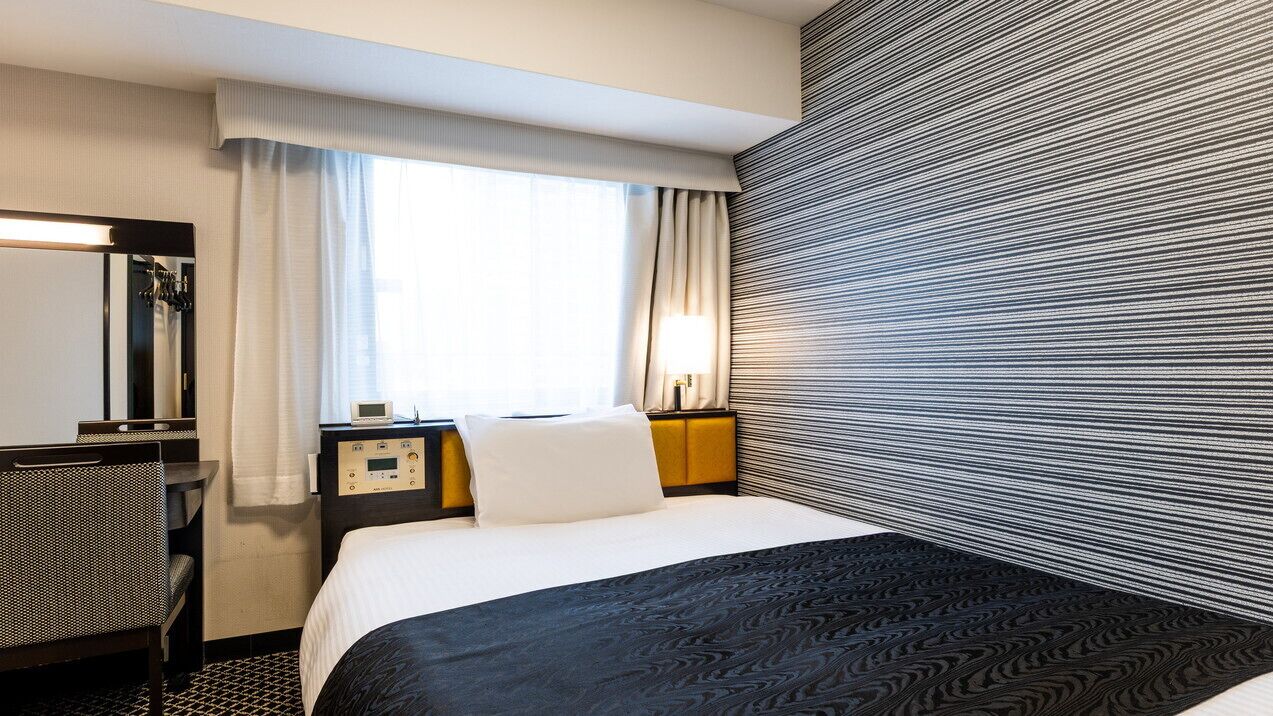 A hotel room with a bed, a desk, a chair, a nightstand, a lamp, and a window with curtains.