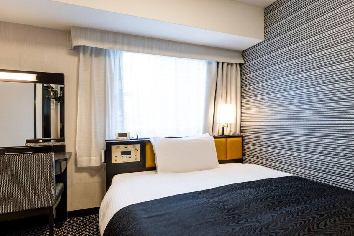 Een hotelkamer met een bed, een bureau, een stoel, een nachtkastje, een lamp en een raam met gordijnen.