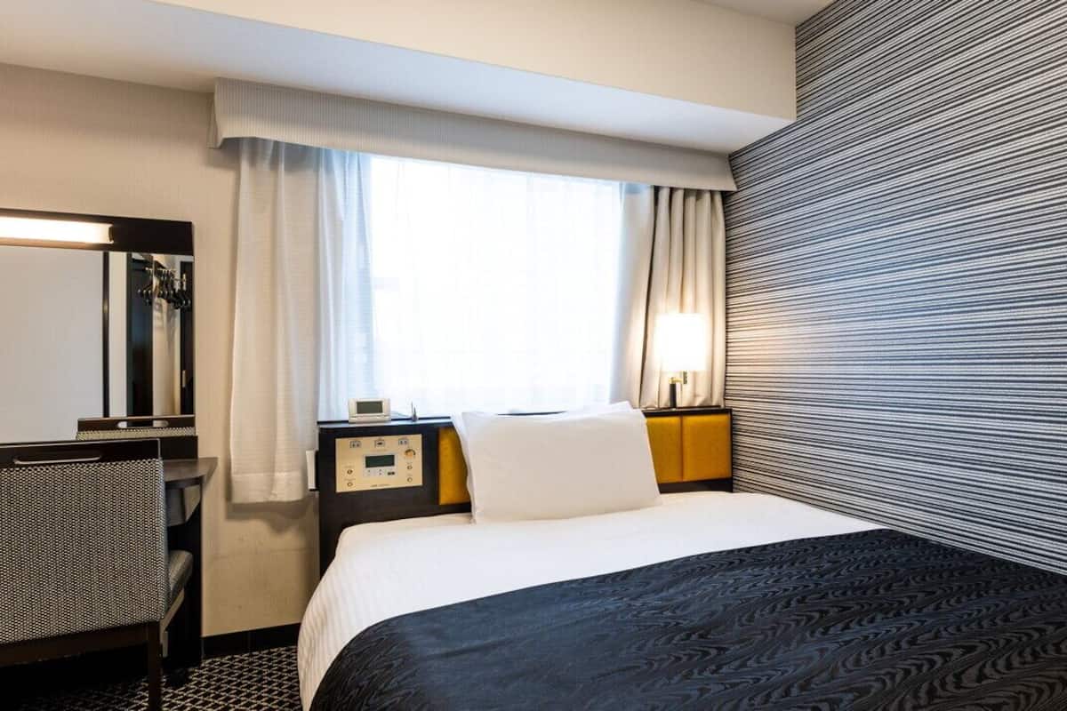 Une chambre d’hôtel comprenant un lit, un bureau, une chaise, une table de chevet, une lampe et une fenêtre avec des rideaux.