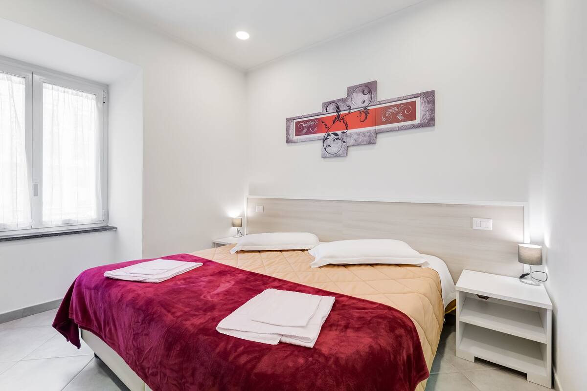 A hotel room with a bed, a nightstand, a wall-mounted artwork, and a window with curtains.
