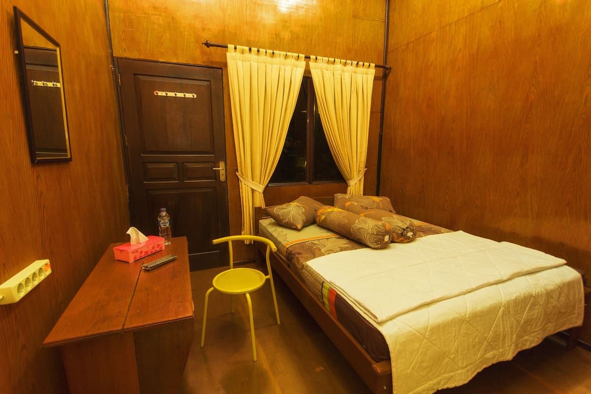 A room with a wooden door, a bed with a white bedspread, a yellow chair, and a small table with a remote control and tissues.