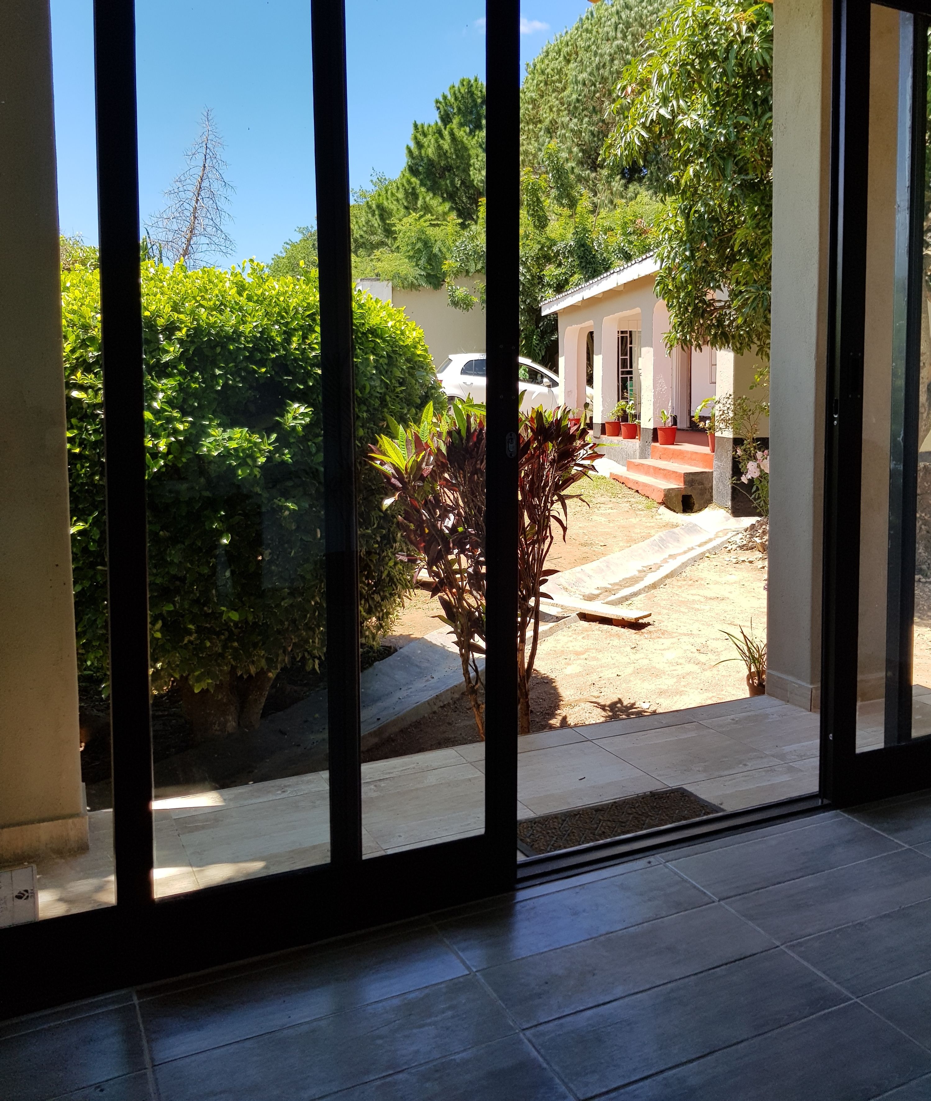 A view through a glass door of a house with a paved driveway, a white car parked outside, and a small garden with potted plants.