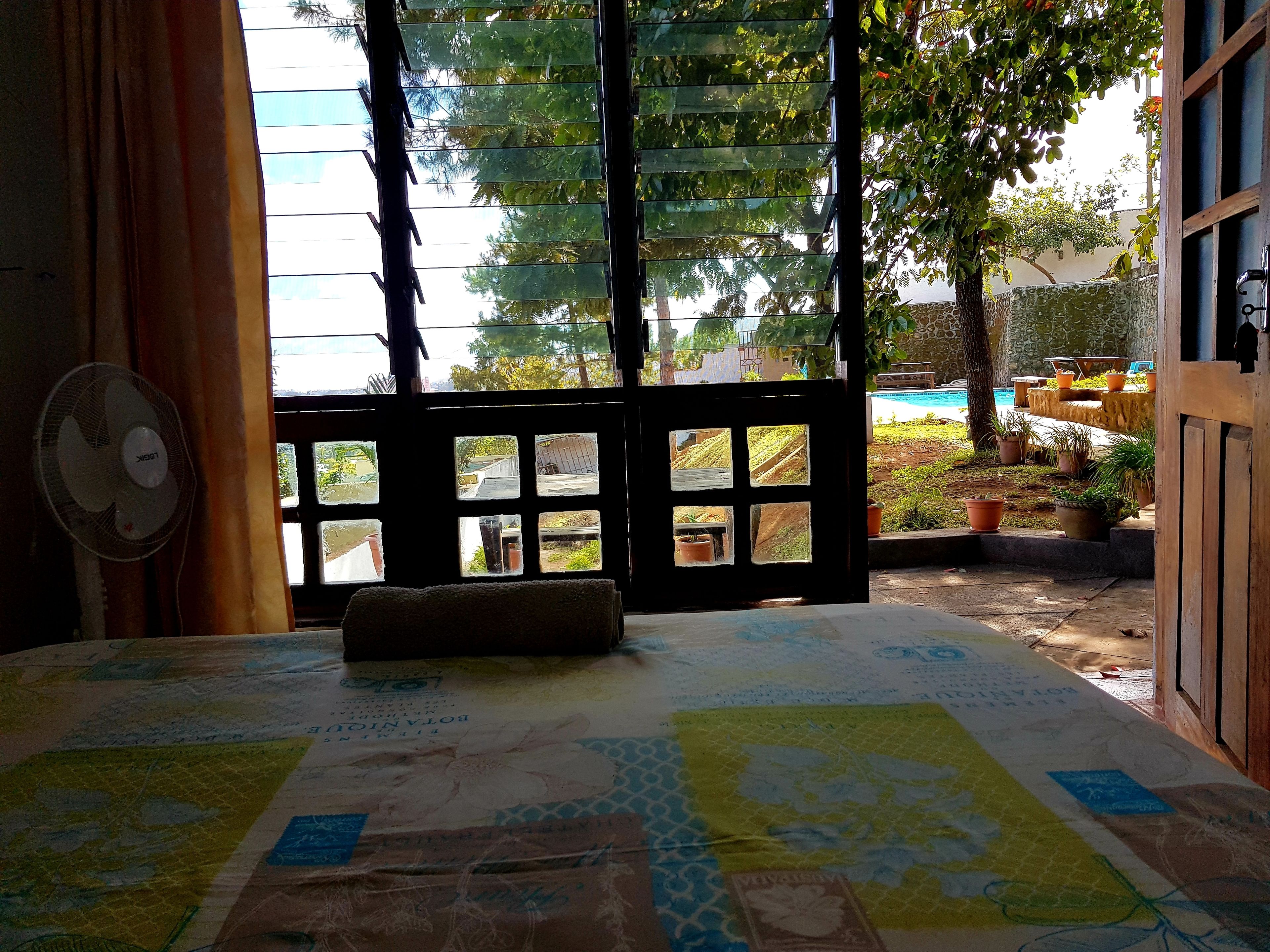 Une chambre avec un lit, un ventilateur et une vue sur un jardin et une piscine, visible par une fenêtre.