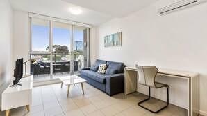 A modern living room with a sofa, a desk, and a chair. There is a TV on a stand, a window with a view of the outside, and an air conditioning unit on the wall. - Poplar Sanctuary Apartments (Box Hill)