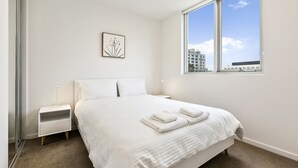 A neatly made bed with white linens and towels, a bedside table with a lamp, and a window with a view of buildings. - Poplar Sanctuary Apartments (Box Hill)