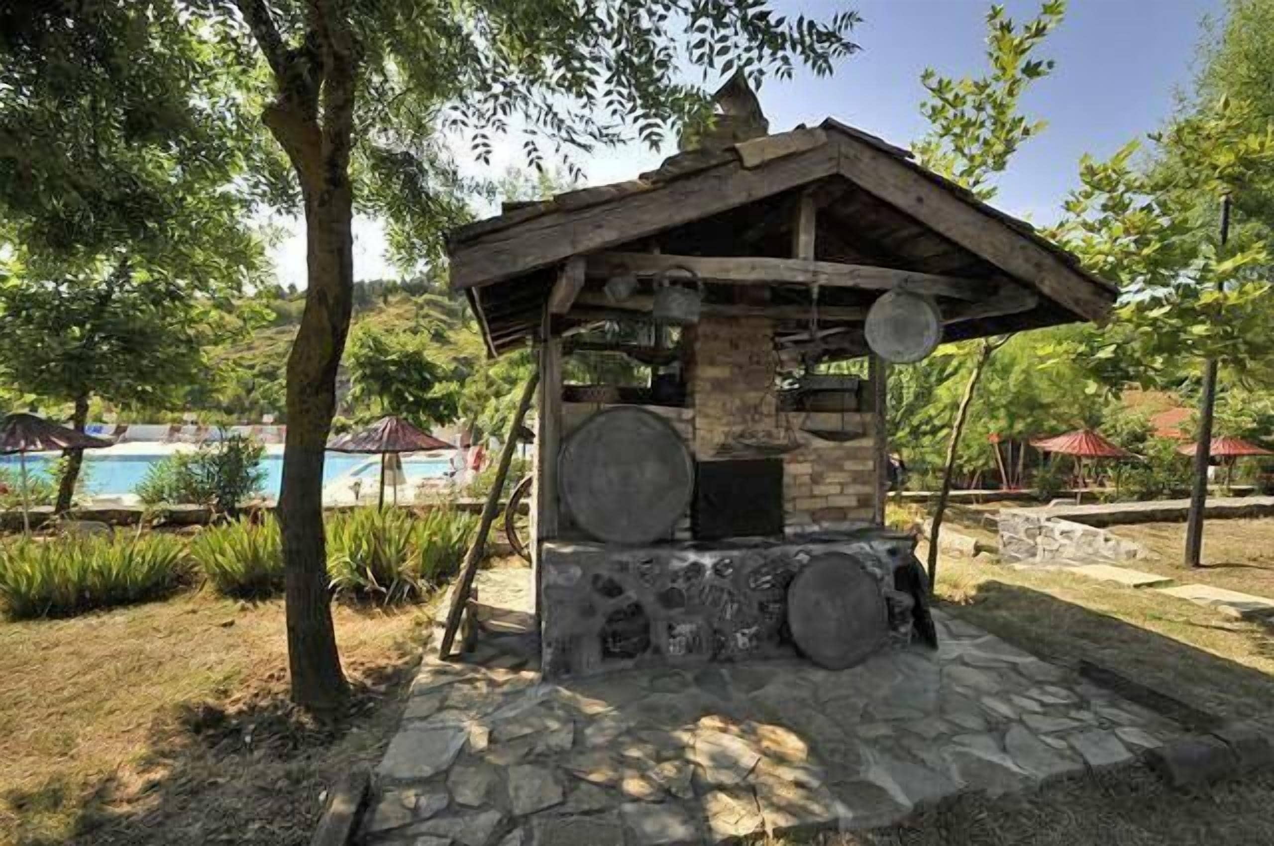 Una estructura rústica al aire libre con base de piedra y techo de madera, rodeada de follaje y una zona de piscina.