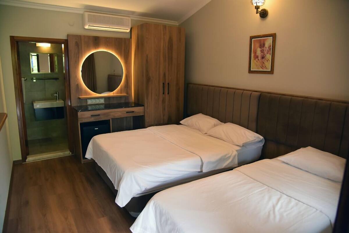 A bedroom with two beds, a wooden wardrobe, a round mirror, and a small bathroom with a sink and mirror.
