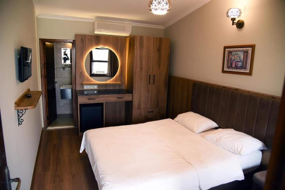 A bedroom with a bed, a wooden wardrobe, a small desk with a mirror, and a wall-mounted light fixture.