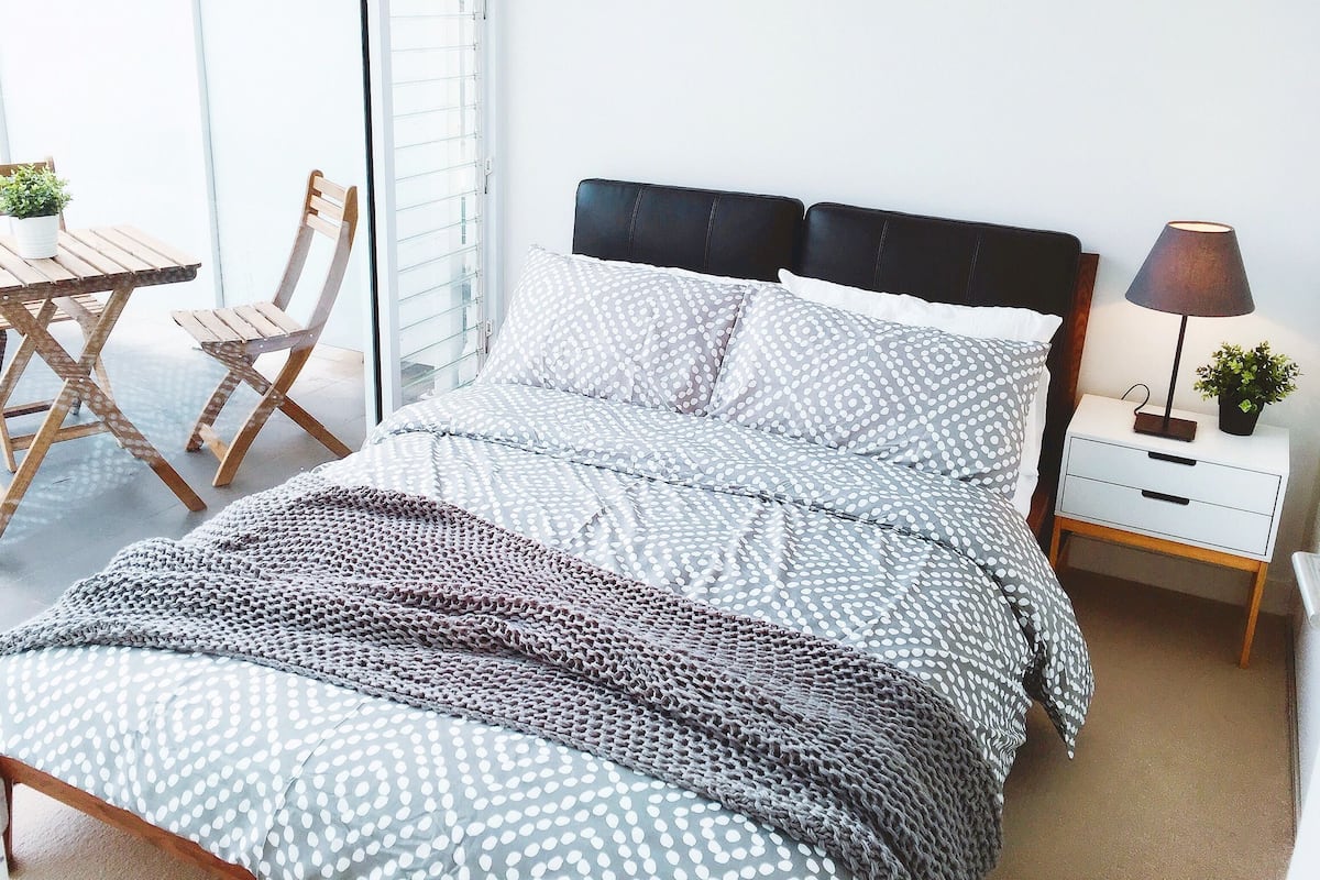A bedroom with a bed, a nightstand, a lamp, a chair, and a table with a potted plant.