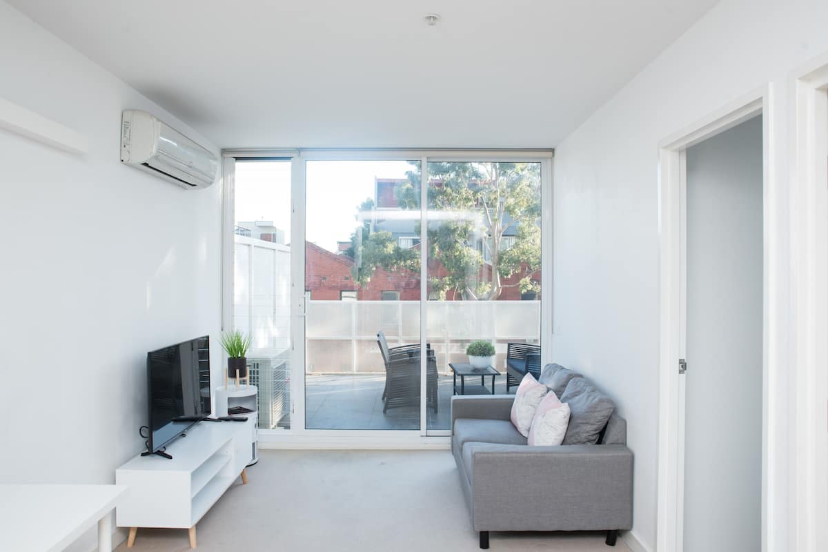 A modern living room with a flat-screen TV, a grey sofa, and a sliding glass door leading to an outdoor patio.