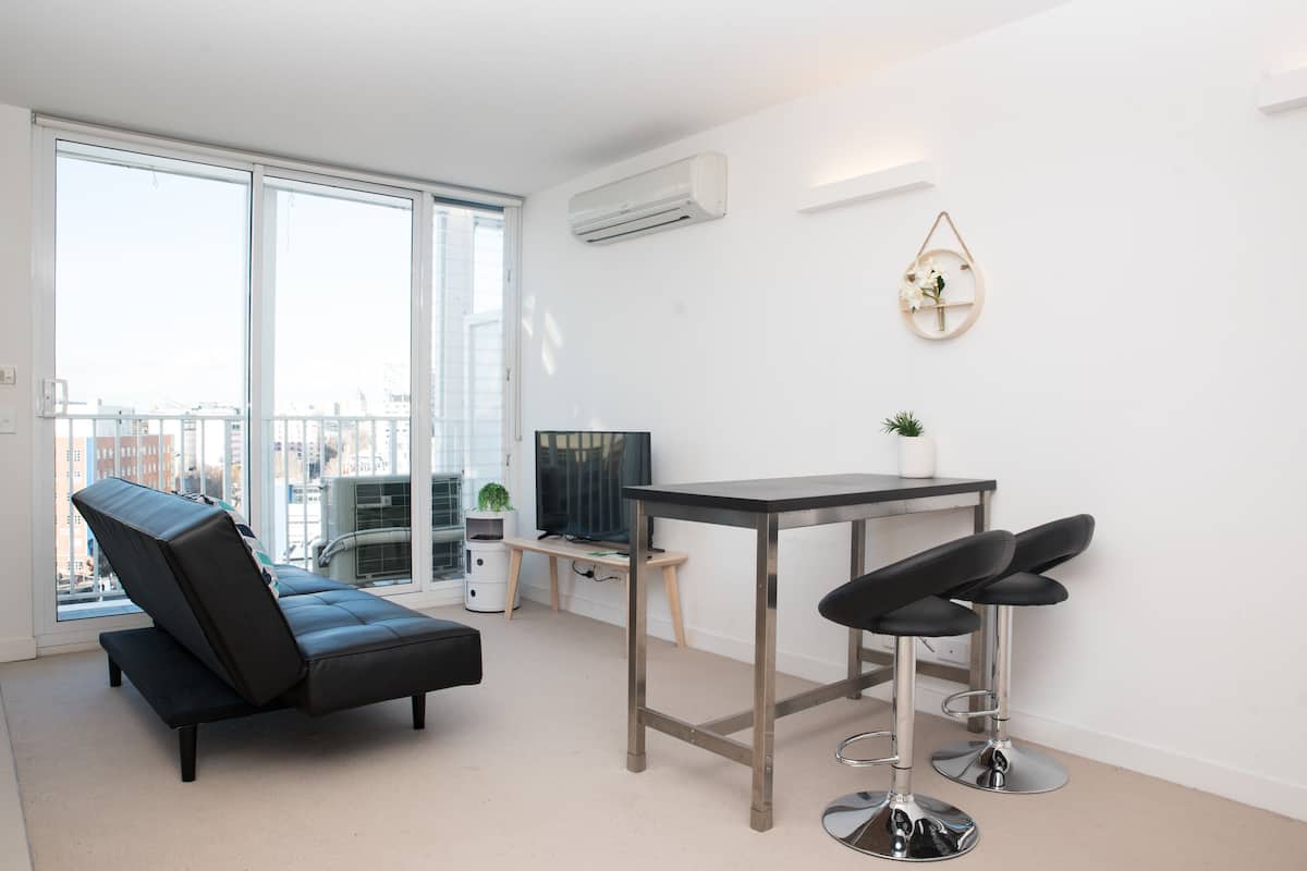 A modern living room with a sofa, a dining table with two bar stools, a TV, and a small plant.