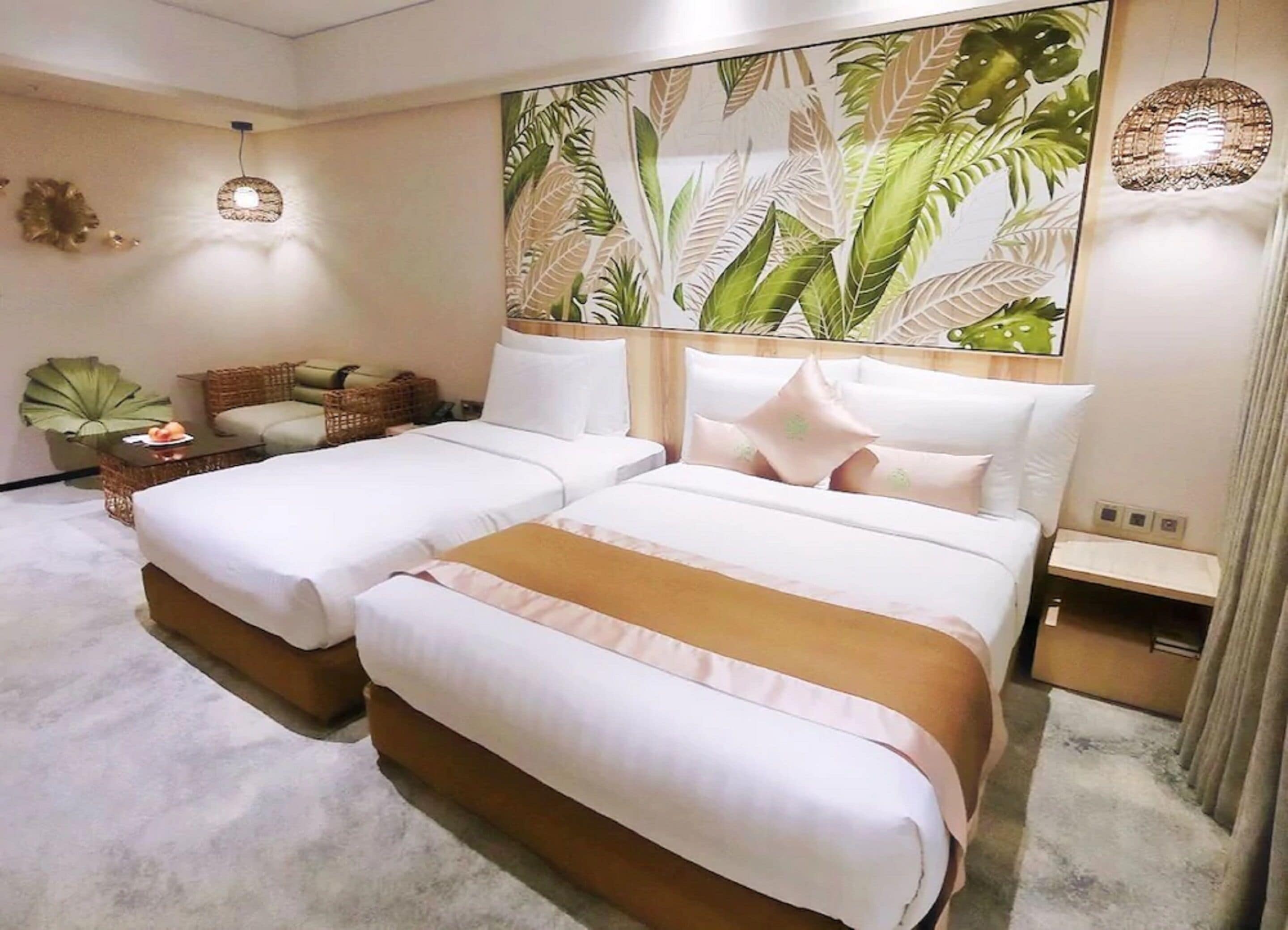 A hotel room with two beds, a large tropical leaf mural, a wicker sofa, and a small table with a lamp.