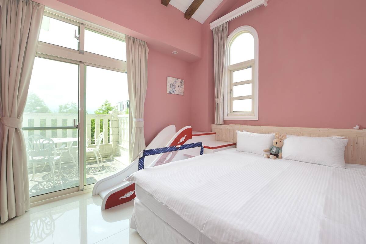 A bedroom with a large bed, a balcony with a table and chairs, and a window with curtains.