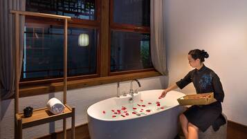 Una mujer sentada en una bañera llena de pétalos de rosa, sosteniendo una caja de rosas rojas.