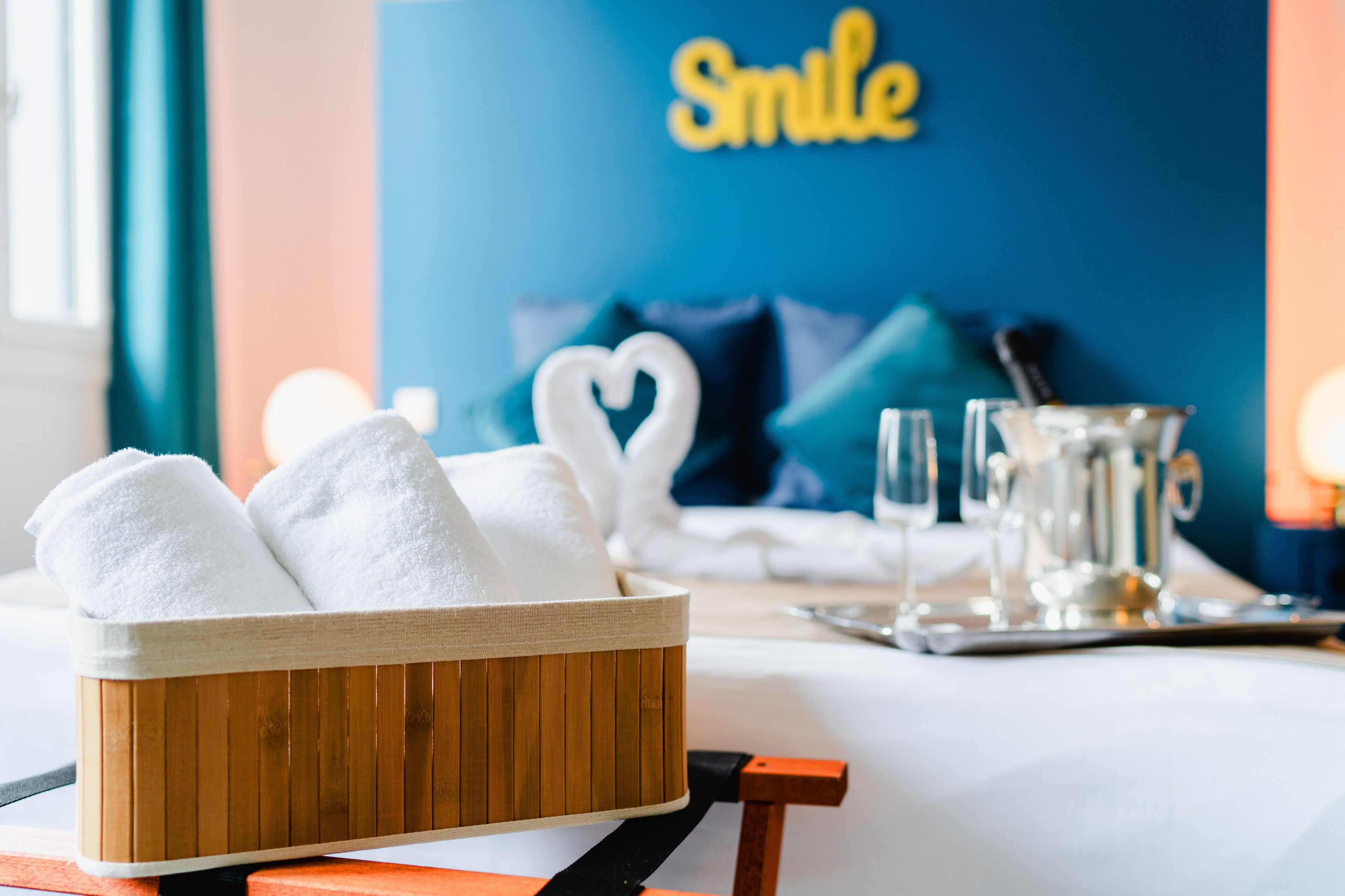 A wooden basket with white towels, a heart-shaped decoration, and a tray with glasses and a pitcher on a table in a room with a blue wall and a 'Smile' sign.