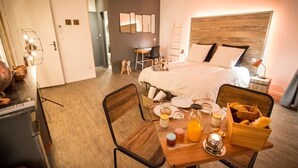 A hotel room with a bed, a wooden dining table with breakfast items, a desk with a chair, and a small side table with a lamp. - La Maison Gustave (Narbonne)