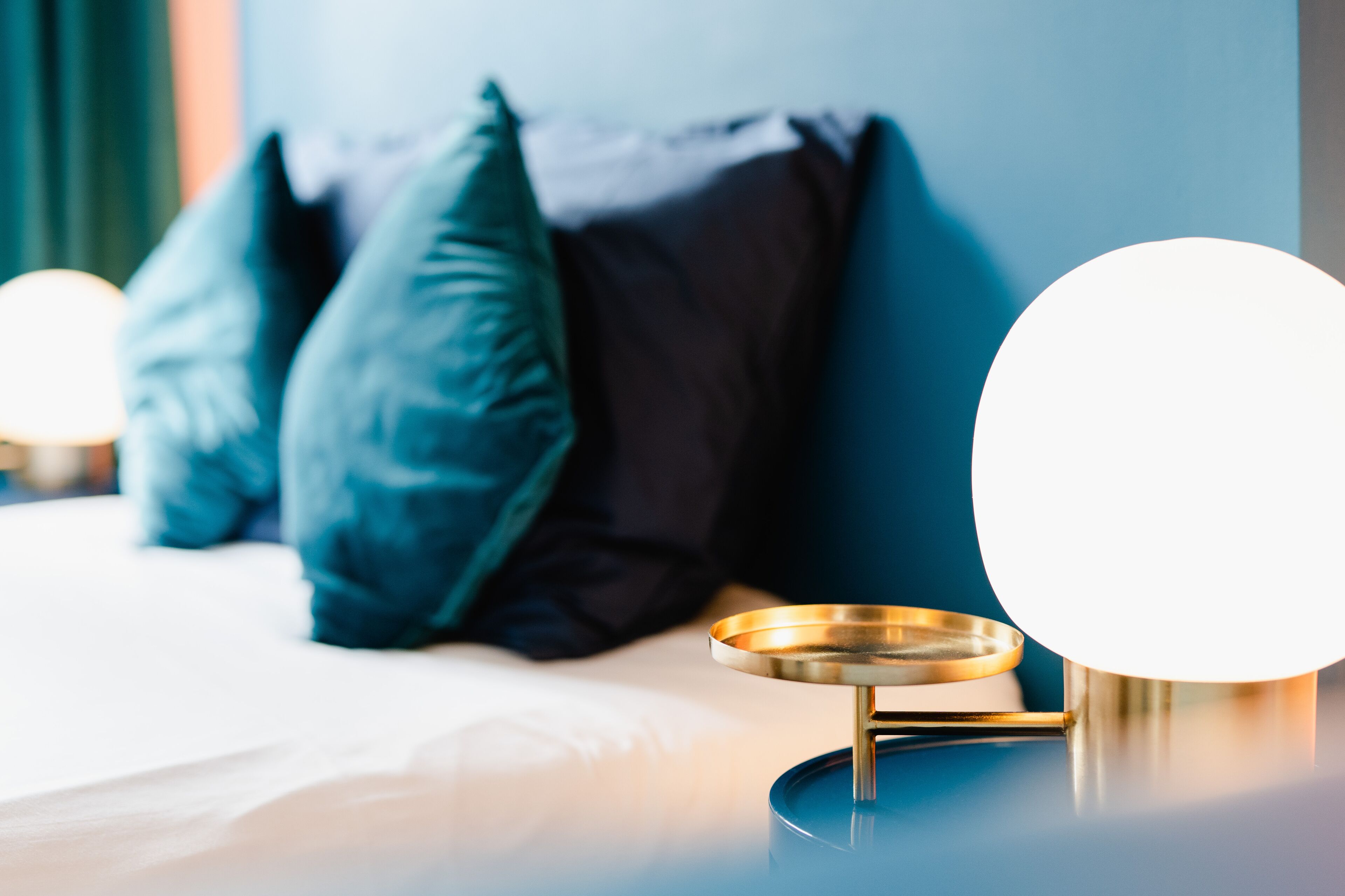 A neatly made bed with pillows, a bedside table with a lamp, and a wall with a circular light fixture.