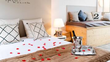 A neatly made bed with a brown quilt, a champagne bottle, and glasses on a tray, along with rose petals scattered on the bed.