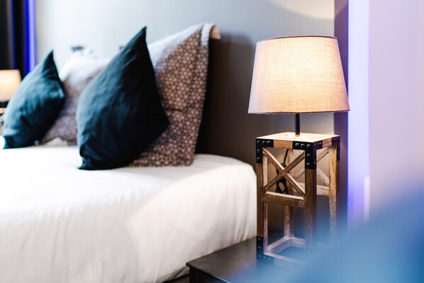 A neatly made bed with dark and patterned pillows, a small wooden side table with a lamp, and a wall with a purple hue. - La Maison Gustave (Narbonne)