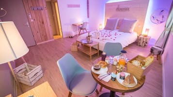 Ein Hotelzimmer mit Holzboden, einem Bett mit Holz Kopfteil, einem Schreibtisch mit Lampe, einem Stuhl, einem kleinen Esstisch mit Frühstücksutensilien und einer Wanduhr.