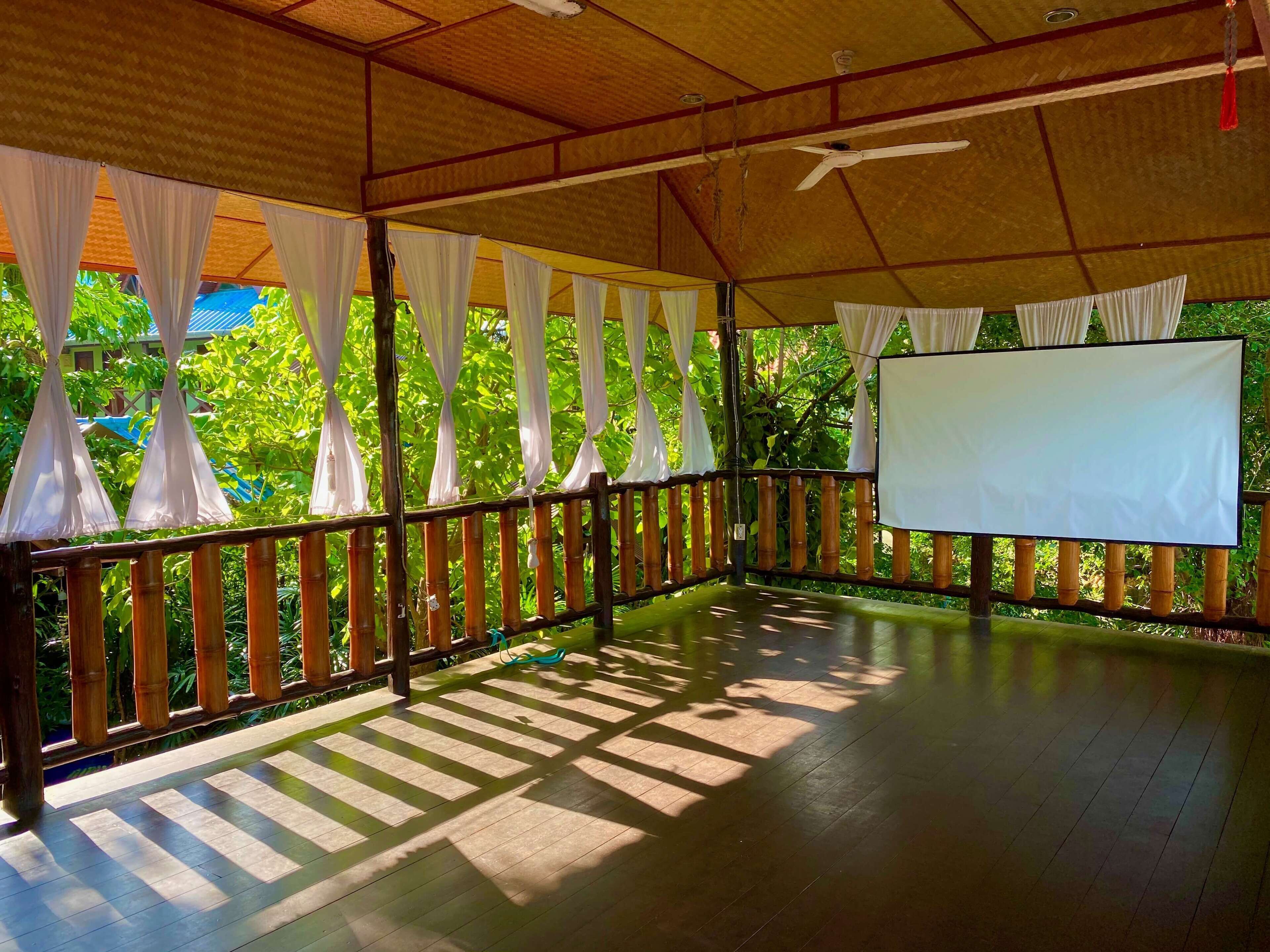 An open-air pavilion with a wooden floor, white curtains, and a blank projection screen.