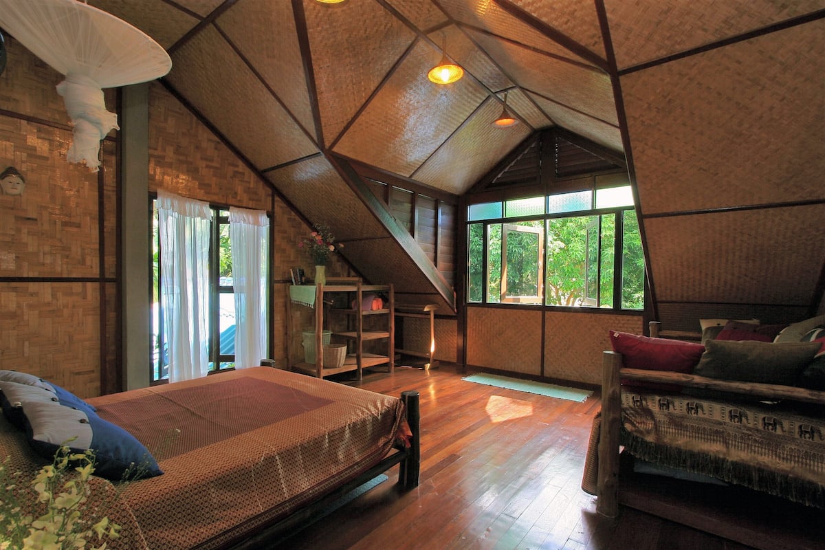 Um quarto amplo com teto alto, vigas de madeira, cama com roupa de cama estampada, sofá com almofadas e janela com vista para o verde lá fora.