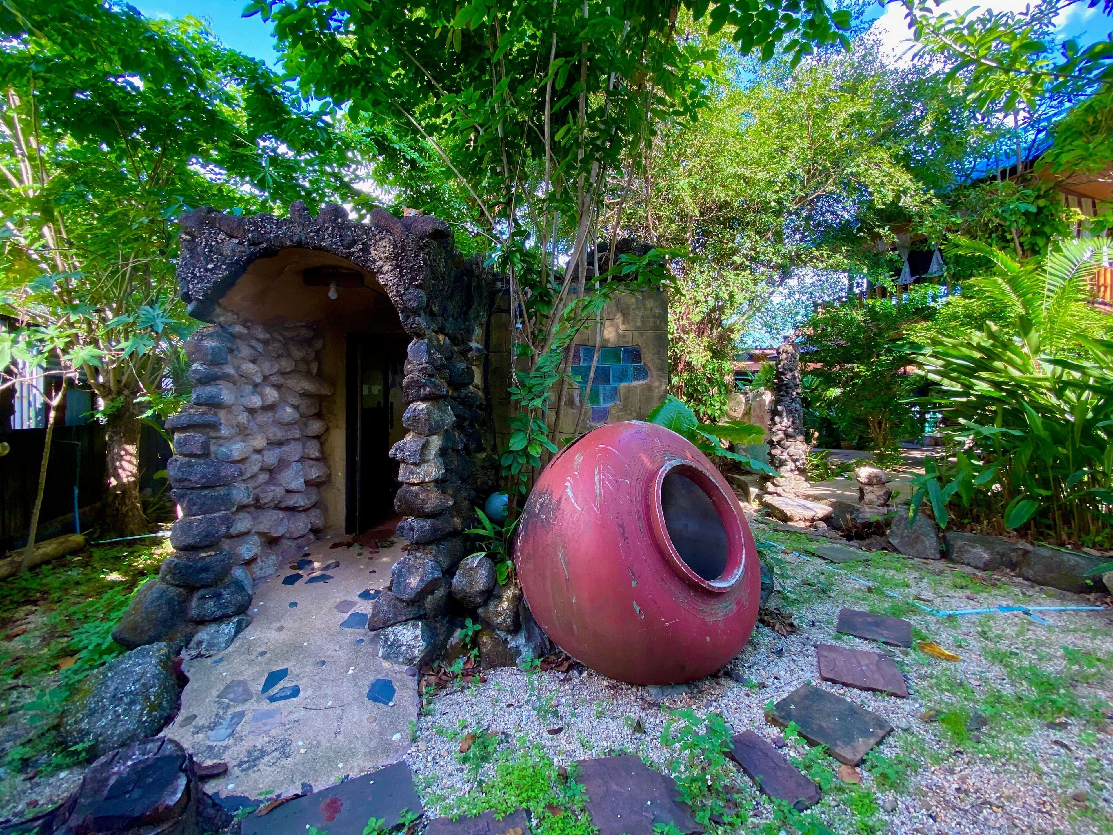 A rustic stone structure with a small entrance, a large red pot, and lush greenery surrounding the area.