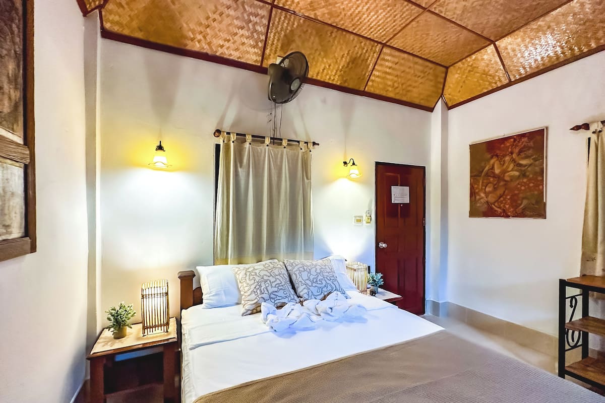 Quarto de hotel com uma cama grande, duas mesas de cabeceira, um ventilador fixado na parede, uma porta com uma placa e um quadro na parede.