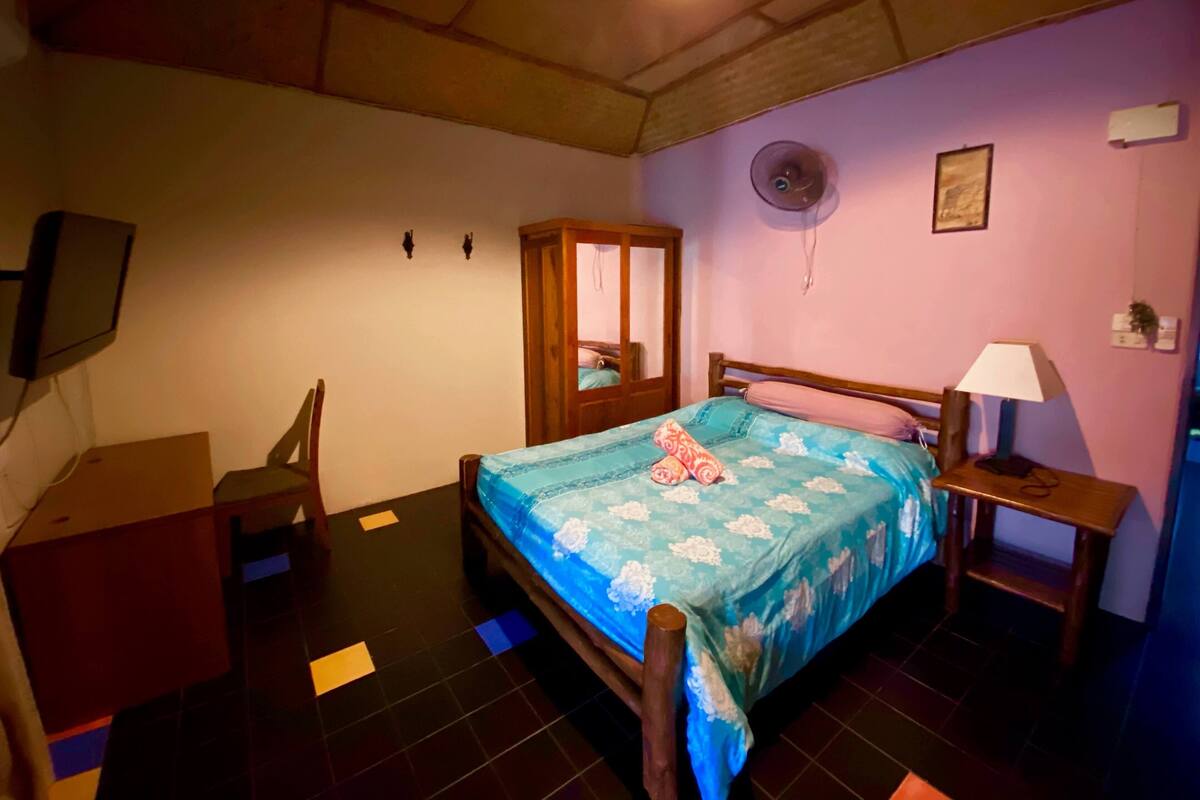 Quarto com cama, guarda-roupa de madeira, escrivaninha, cadeira, mesa de cabeceira com abajur e ventoinha fixada na parede.