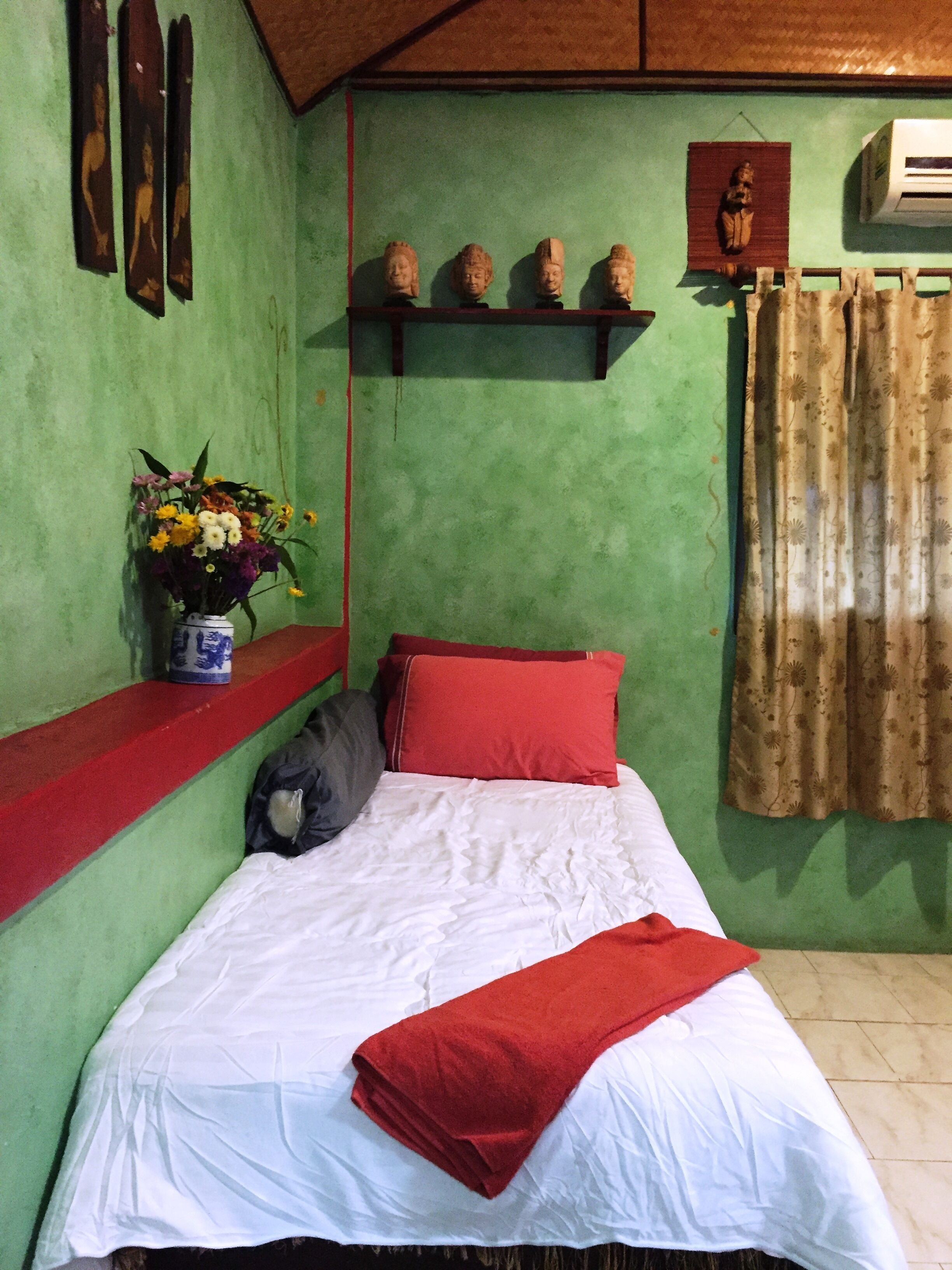 Une pièce comprenant un lit, un oreiller rouge, un drap blanc, une couverture rouge, un vase avec des fleurs et une étagère murale ornée d’objets décoratifs.