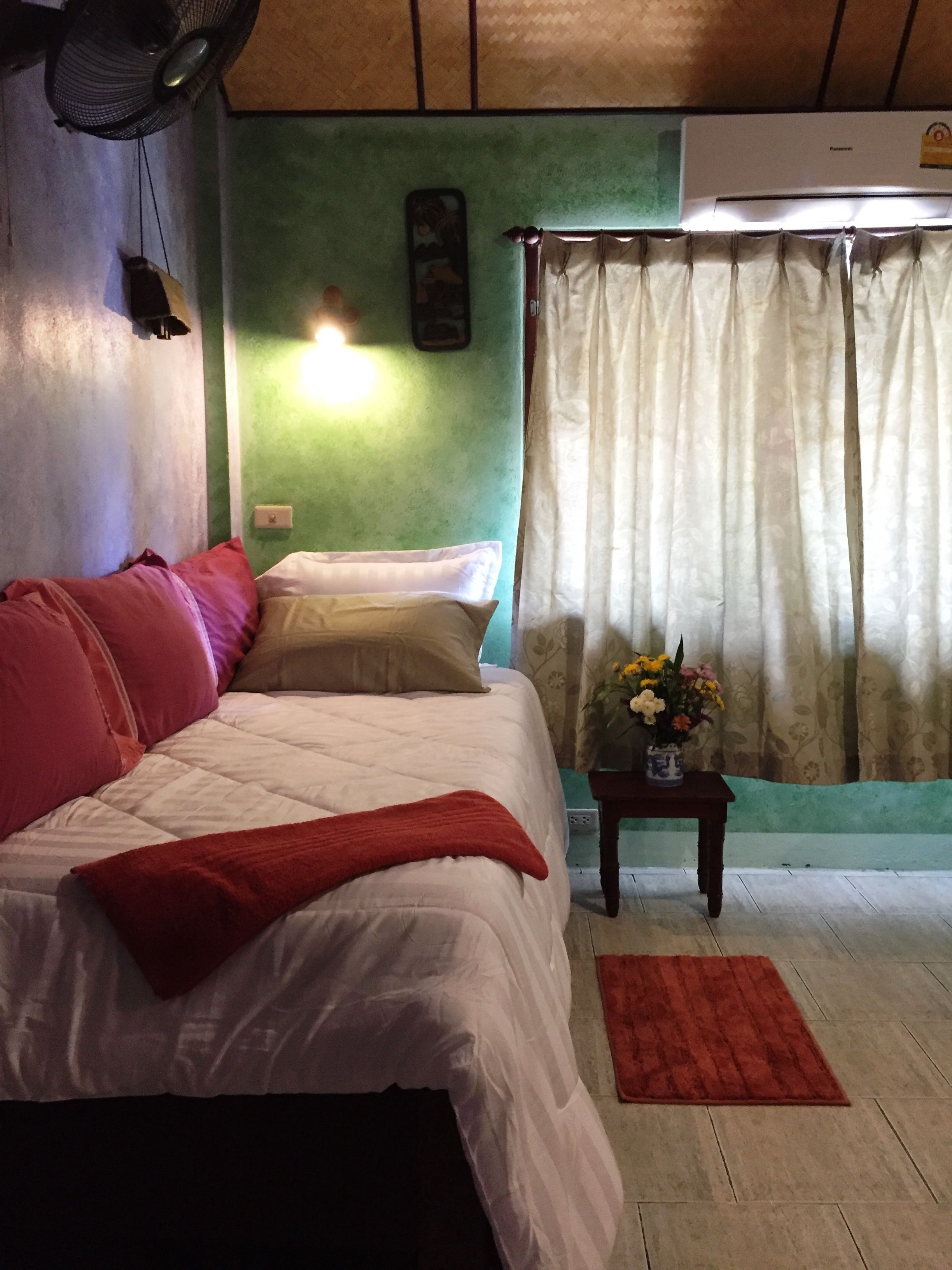 A bedroom with a bed, pillows, a bedside table with a vase of flowers, a fan, and a curtain.