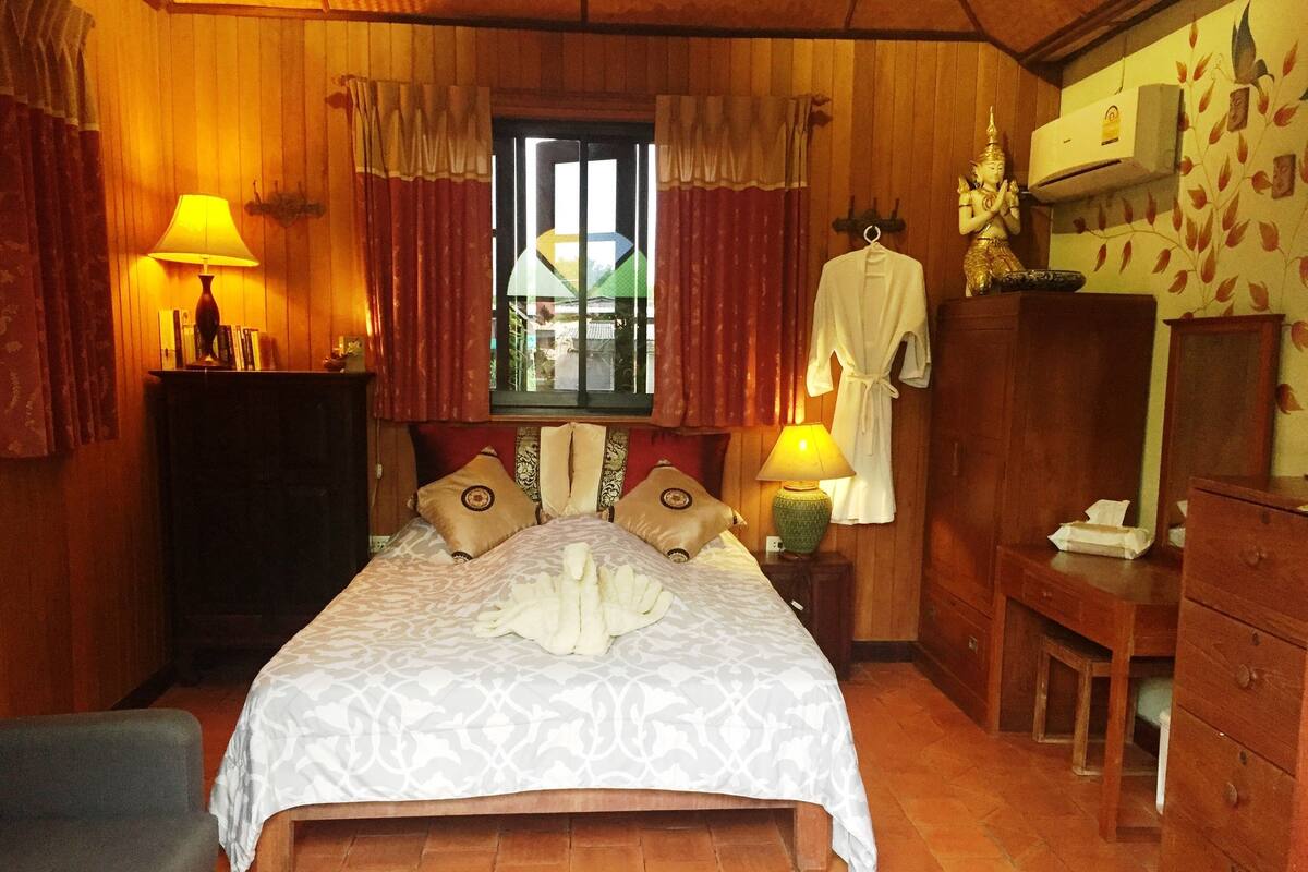 Een goed verlichte slaapkamer met een bed, kussens, een nachtkastje, een lamp, een badjas, een spiegel en een kledingkast.