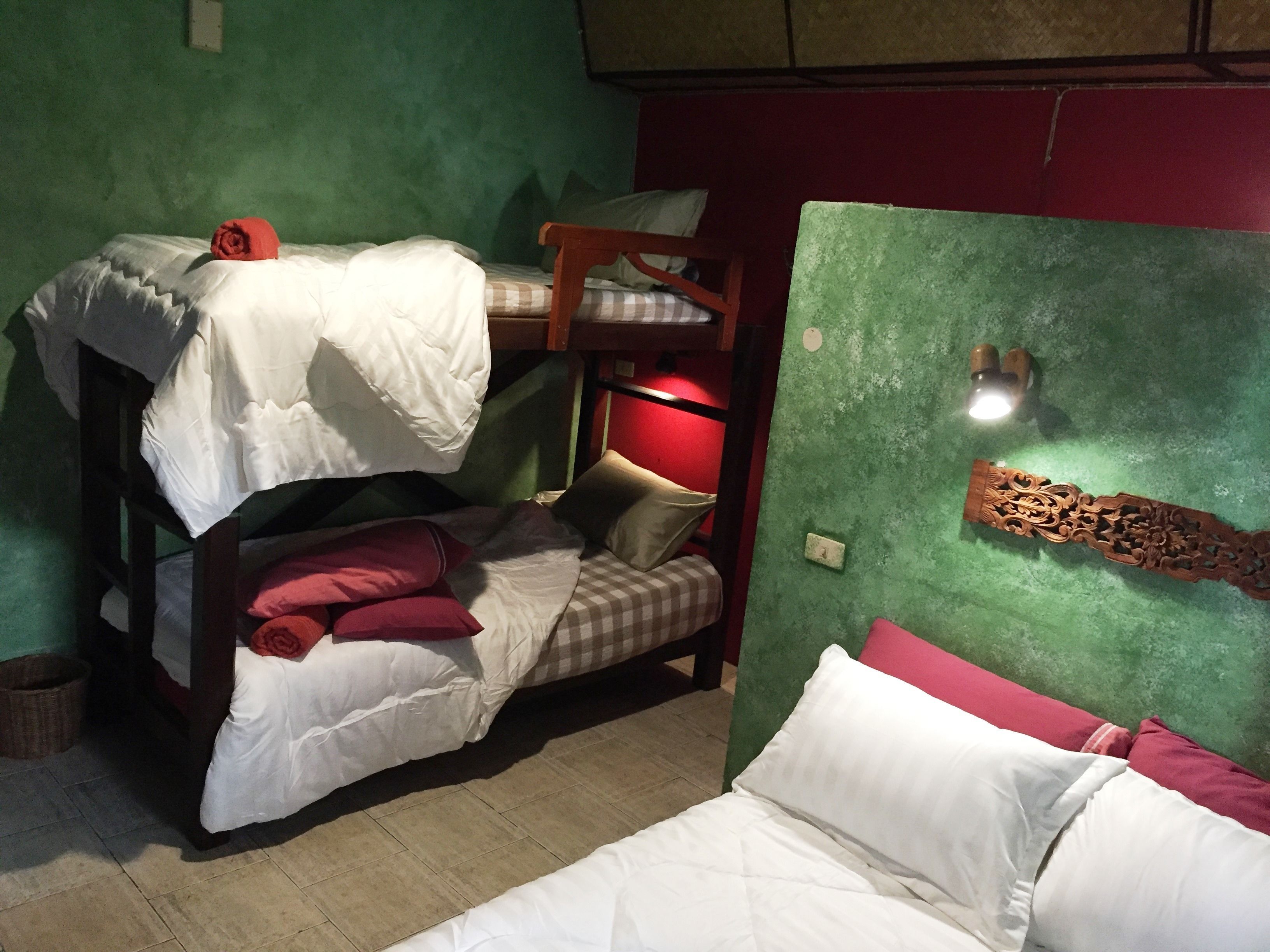 A bunk bed room with a green wall, a red wall, and a wooden headboard. There is a small table with a lamp and a power outlet on the green wall.