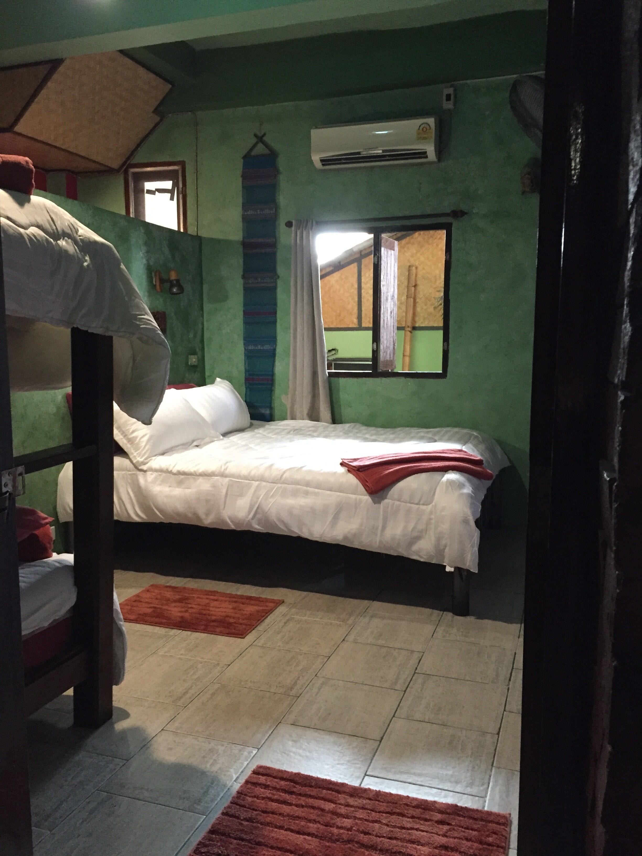 A room with a bunk bed, a single bed, a window with curtains, an air conditioning unit, and a red rug on the floor.