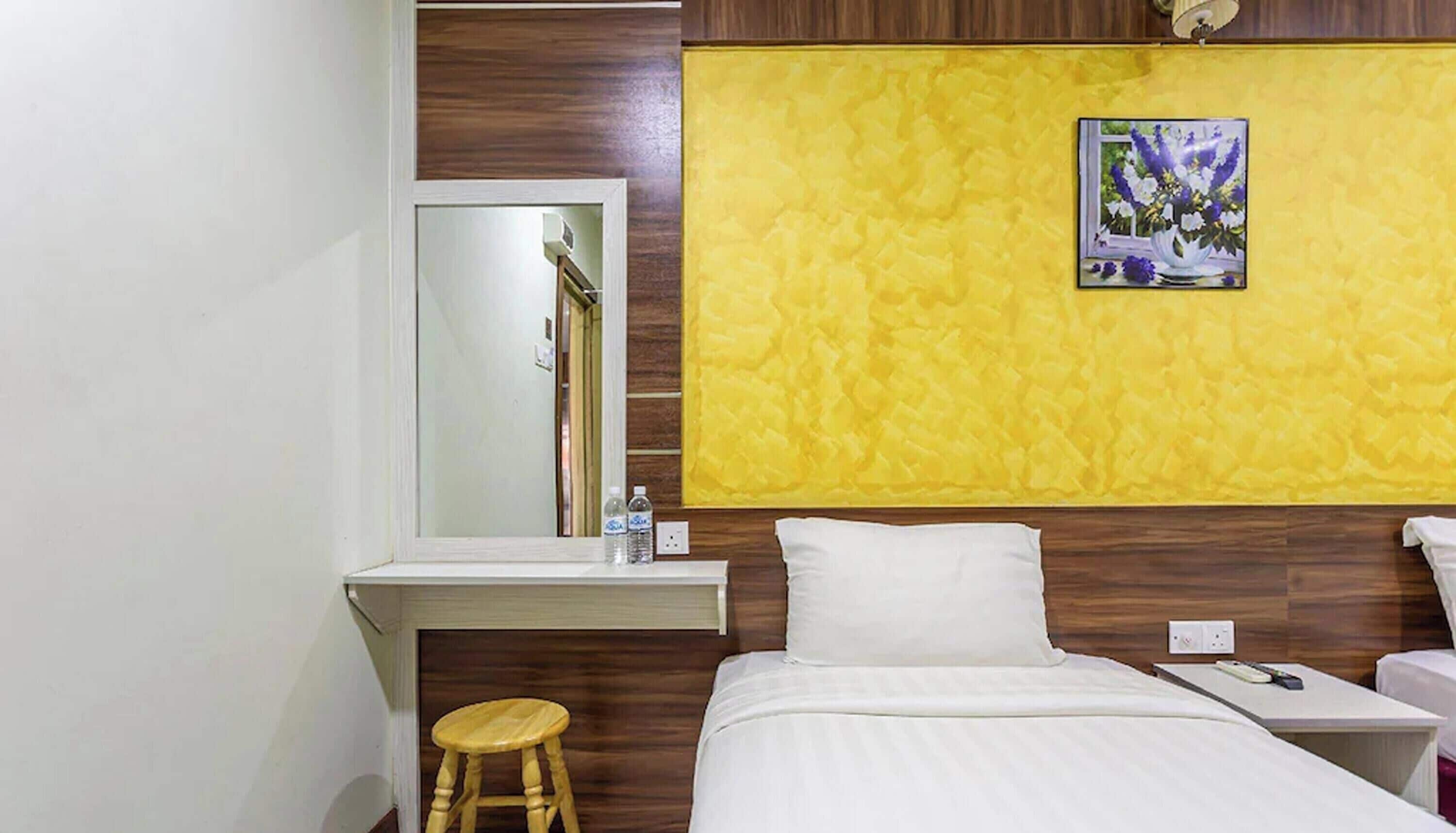 A hotel room with a bed, a bedside table, a mirror, a stool, a yellow textured wall, and a framed picture.