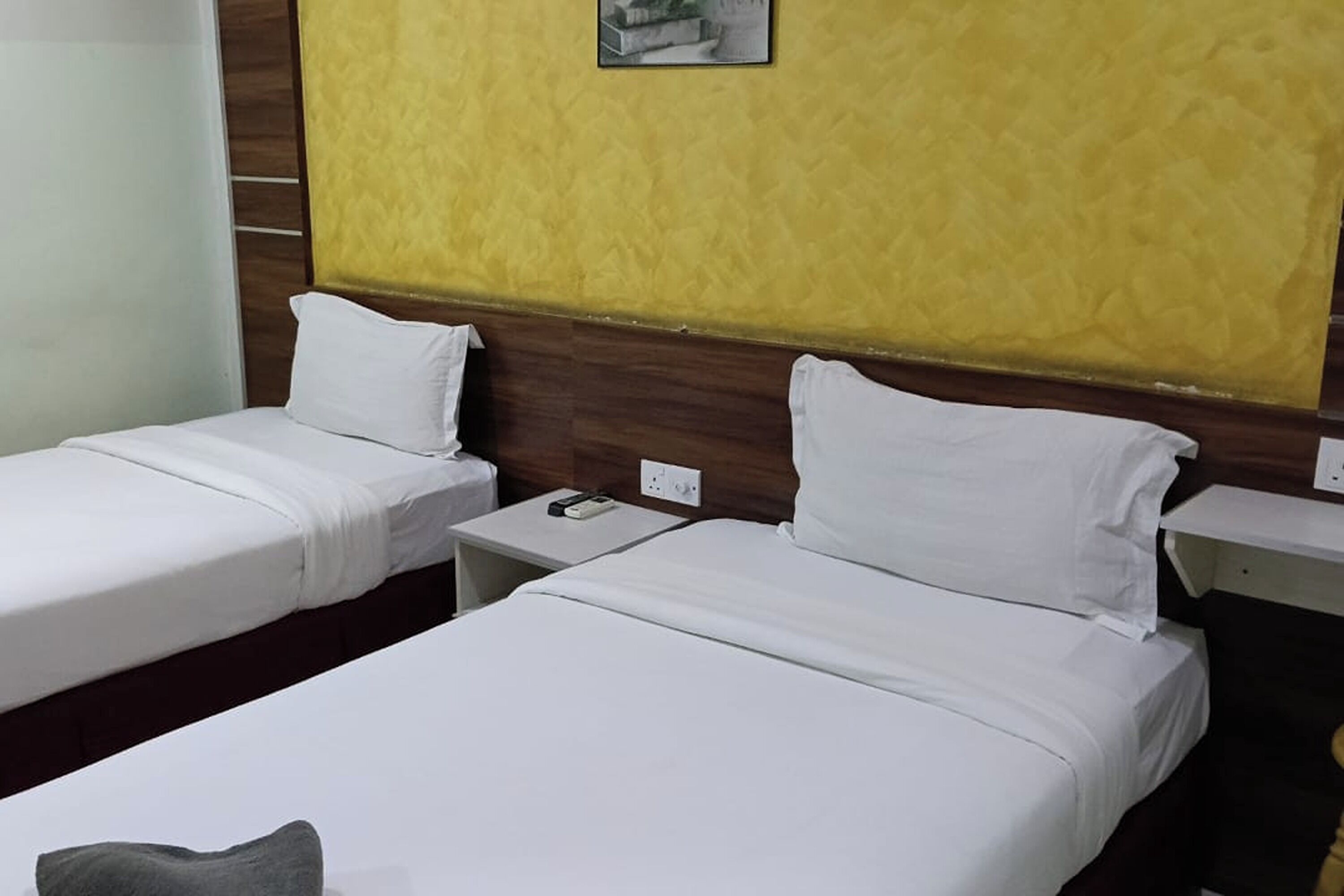 Habitación de hotel con dos camas individuales, un cabecero de madera, una mesita de noche pequeña con un teléfono y un interruptor de luz en la pared.